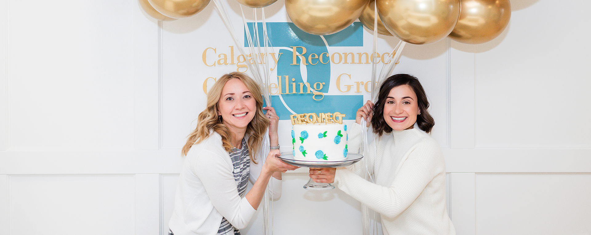 Two psychologists holding a cake decorated with gold letters spelling RECONNECT, stand in front of a logo sign that says Calgary Reconnect Counselling Group. Gold balloons float above in the background.