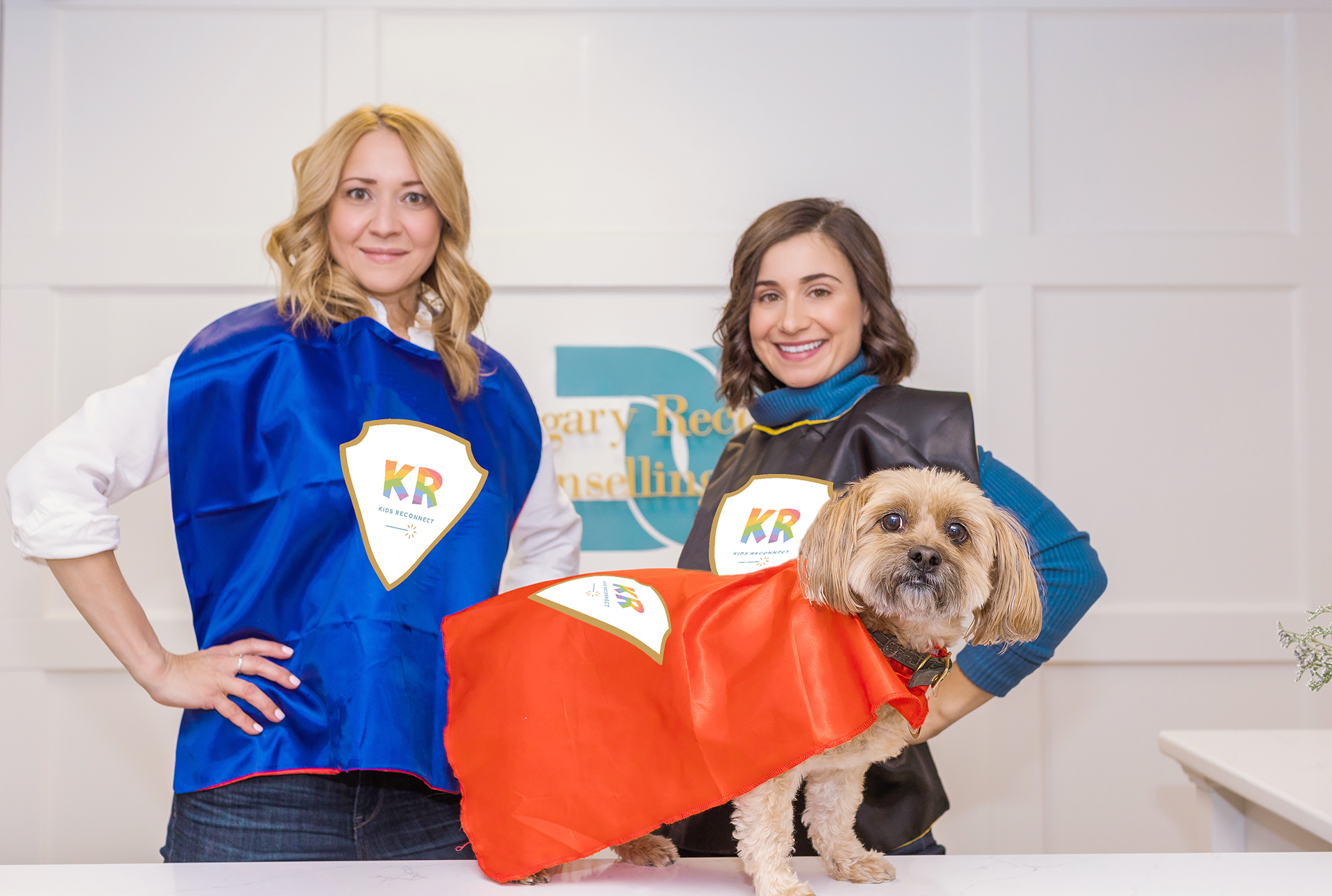 CRCG clnic directors Natalie Bergman and Marcella Galizia, and a small dog, wearing Superhero capes with the Kids Reconnect logo.