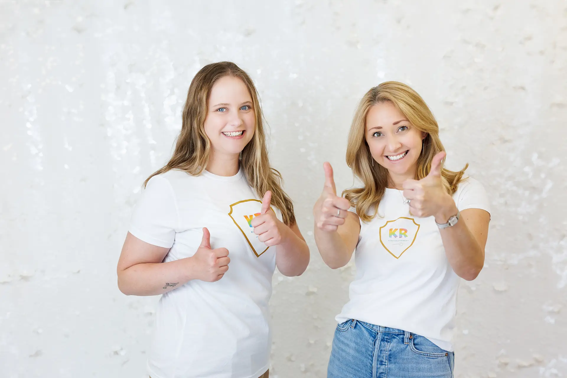 Two women smile as they stand side by side, making a thumbs up gesture with both hands.